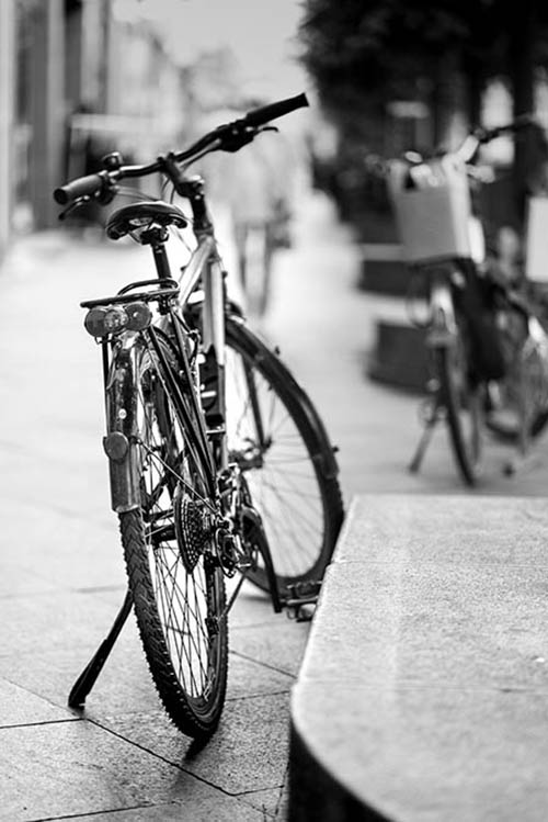 black and white image of an old and simple bike, that is stationary, with another stationary cycle in the background that is slightly blurred